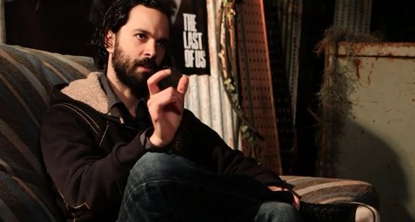 Neil Druckmann menggunakan jaket warna hitam sedang duduk dengan satu kaki terangkat dan sedang mengangkat tangannya